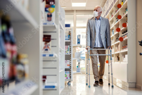 Man with walkers for the elderly goes around the shelves with medicines in the pharmacy. An old man with silver hair walks through a pharmacy and buys therapy drugs. Face mask, corona virus protection