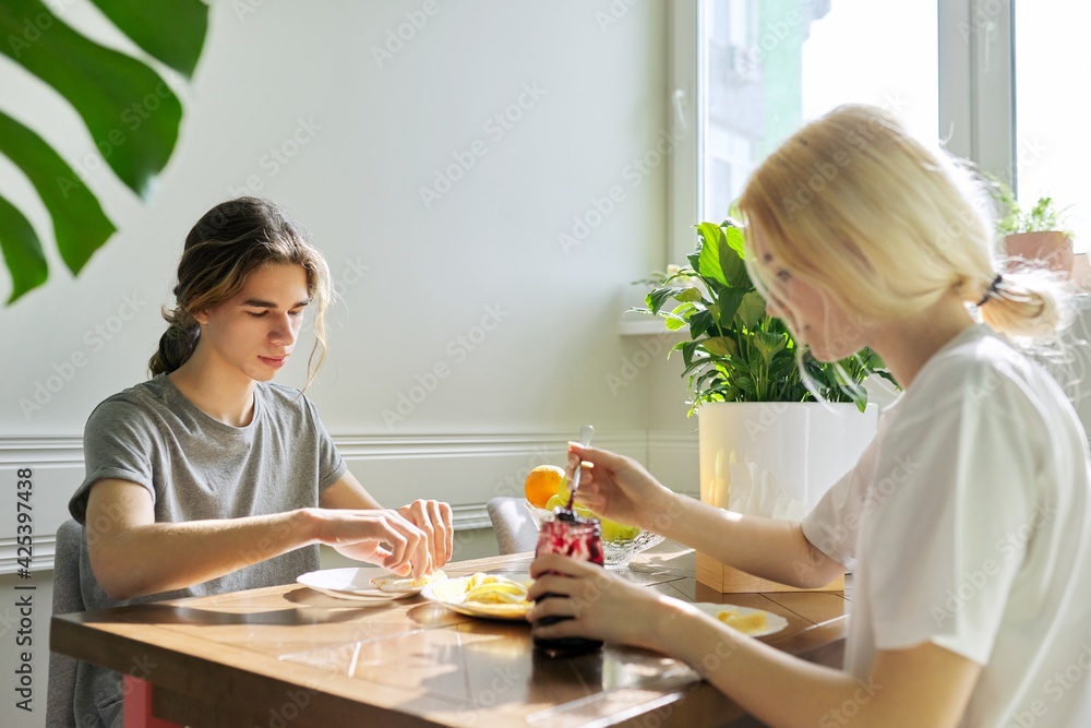 Teenagers guy and girl eating pancakes with jam, sitting at table at home Teenagers guy and girl eating pancakes with jam, sitting at table at home