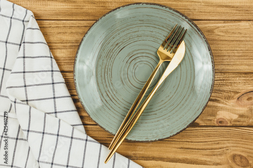 Green empty plate with fork, knife white striped textile napkin on wooden table. Golden cutlery, table setting utensils. 