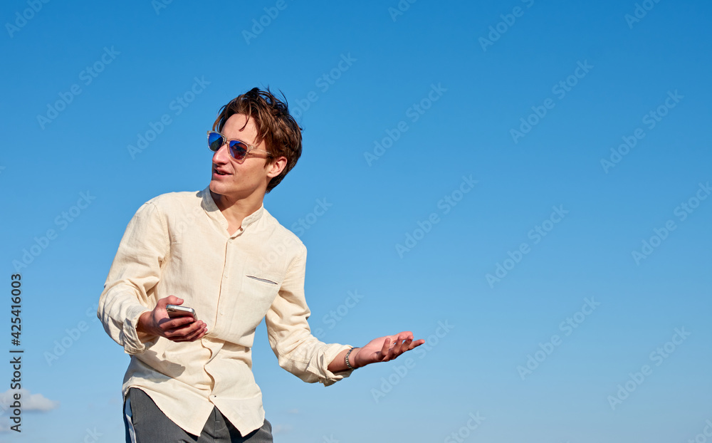 A Spanish white man with long hair, purple glasses and a beige shirt acting confused while holding his phone on clear sky background