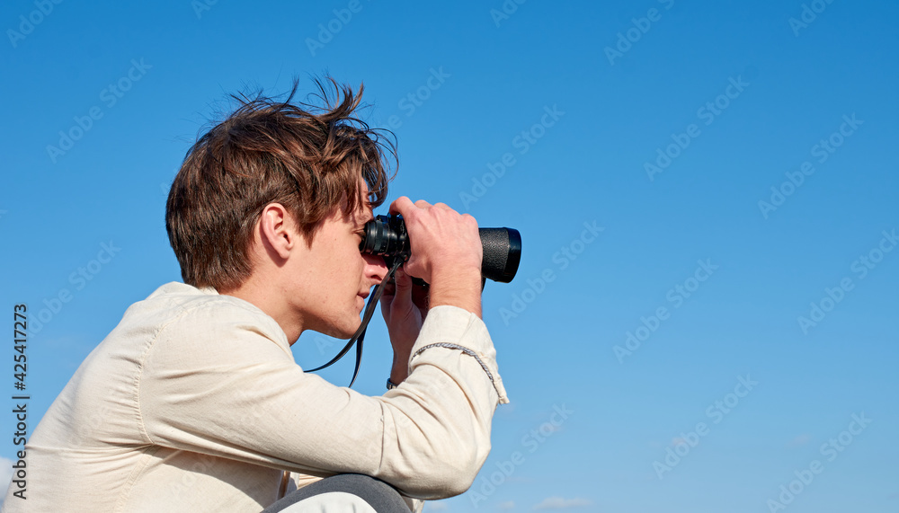 A Spanish white man in a beige shirt looking through binoculars on clear blue sky background
