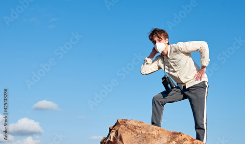 An attractive young man from Spain in a white shirt wearing a mask with binoculars hanging around his neck standing on a cliff, looking afar puzzled