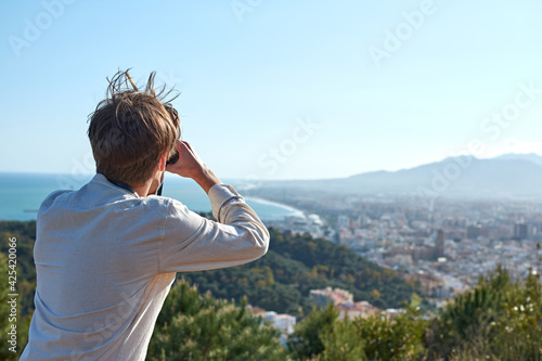 A close up of a young man from Spain in a white shirt wearing a mask observing the coastal town through binoculars