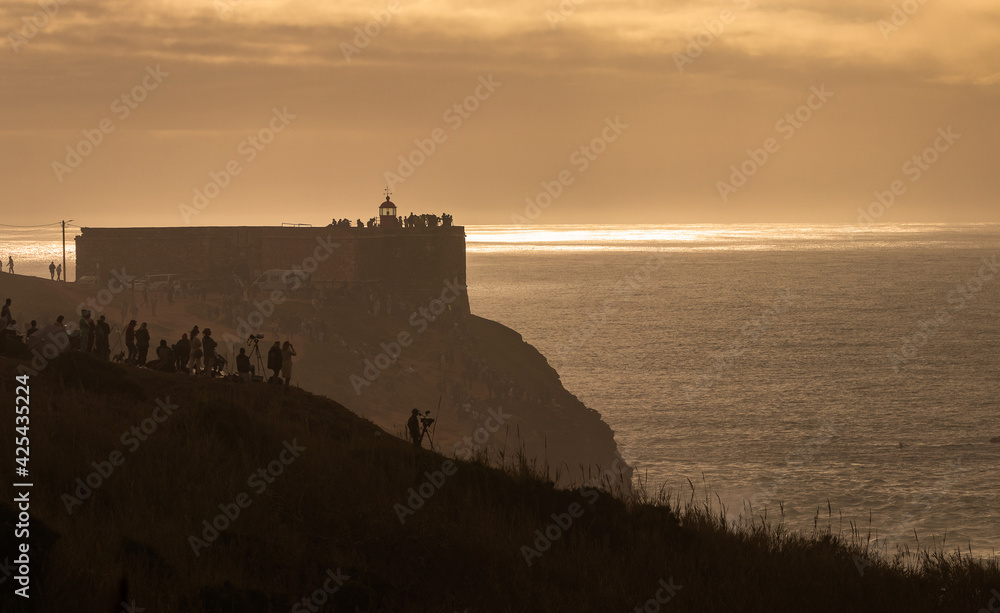 Fototapeta premium Fort next to the North beach in Nazaré, Portugal, seen in silhouette with people in the foreground watching the giant waves and in the background the sunlight mirrored in the sea water.