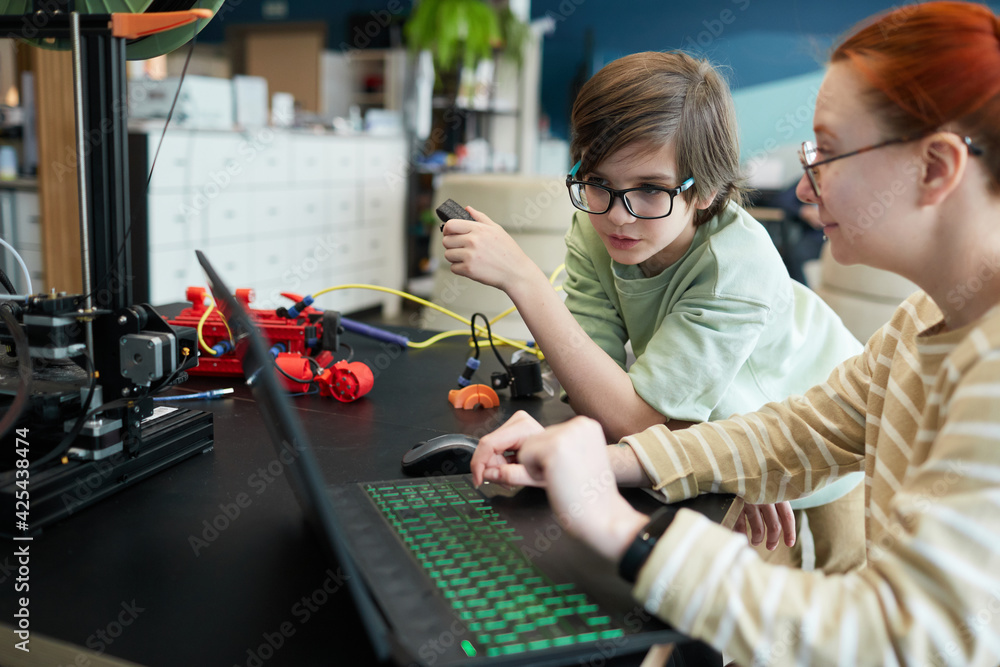 Side view at young female teacher helping boy using 3D printer during robotics and engineering class at school