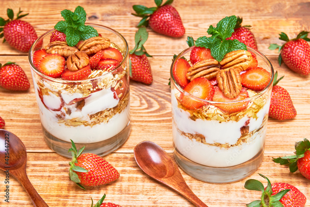 A couple of glasses of strawberry parfait with oat flakes, honey and pecans with a mint leaf on top. on one side whole strawberries and wooden spoons on a wooden table. A couple of glasses of strawberry parfait with oat flakes, honey and pecans with a mint leaf on top. on one side whole strawberries and wooden spoons on a wooden table.