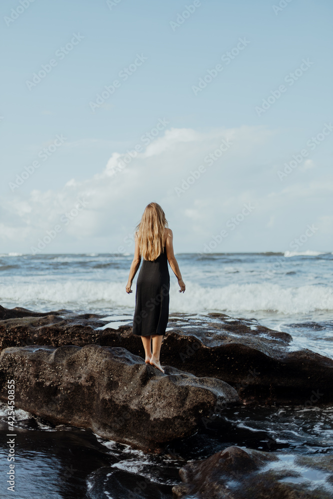 Photoshoot of a blonde girl in a dark dress on the Bali beach with black sand. photo by the cliff, photo by the big stones