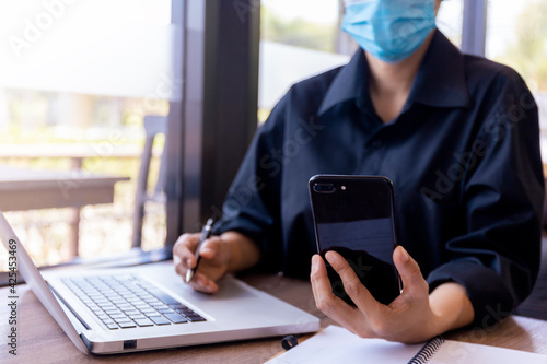 Young woman with protective face mask sitting at coffee shop using smartphone.and laptop. Girl browsing internet, chatting, blogging. Female holding phone and looking on screen. covid-19 new normal