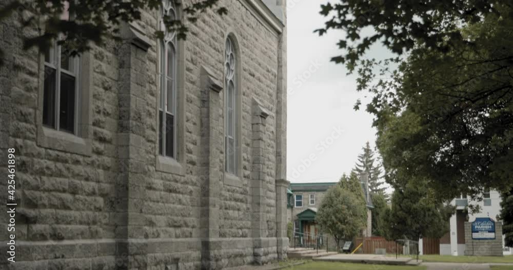 Walking down a concrete path in the middle of the lawn beside the exterior of the building panning up to reveal the steeple of the St. Columban's Church in Cornwall, Ontario, Canada.