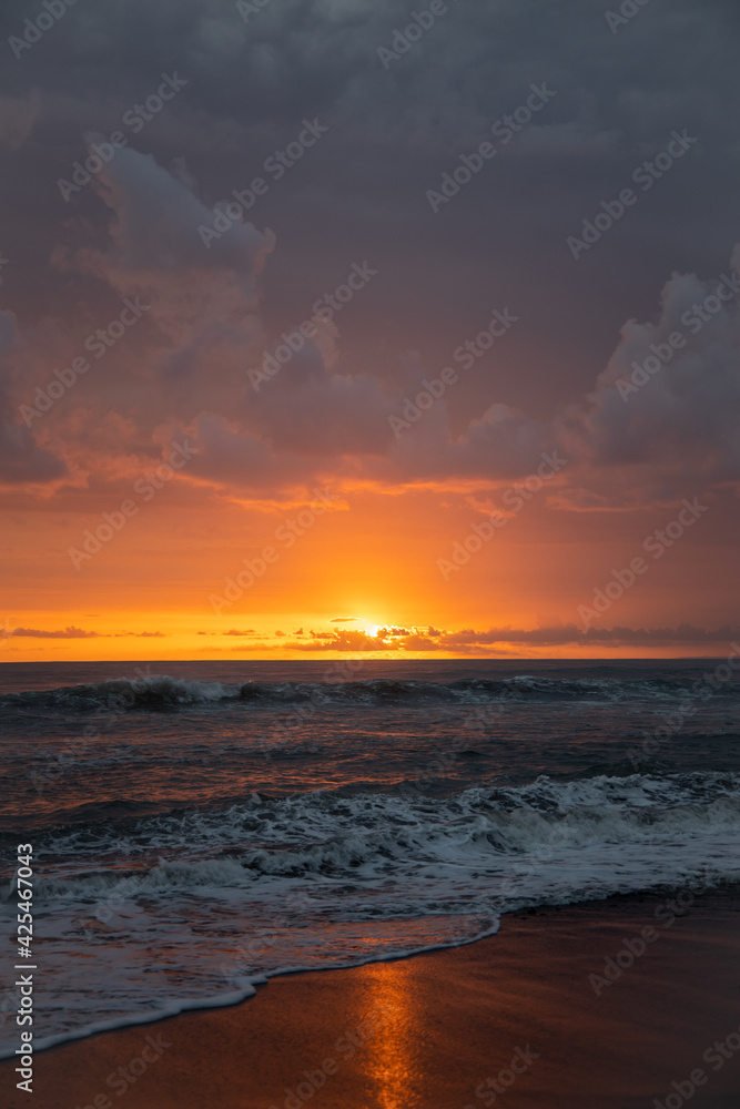 Incredibly beautiful sunset by the ocean in Bali. The bright sun sets in the water. The rays of the sun peeping out from behind the clouds and a beautiful reflection of the sunset in the water