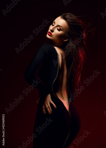Sexy brunette woman vamp stands sideways in black tight unzipped dress demonstrating her naked back, waistline and butt over black background . Fashion, vogue, sexy stylish look for woman concept