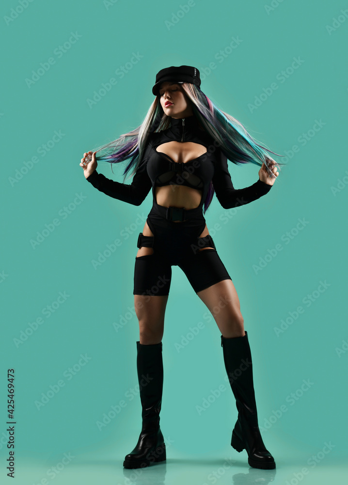 Young blonde woman in black sexy jumpsuit overall, high leather boots and cap stands holding her colored strands in hands over green background, copy space. Sexual games, bdsm, glamour concept Young blonde woman in black sexy jumpsuit overall, high leather boots and cap stands holding her colored strands in hands over green background, copy space. Sexual games, bdsm, glamour concept