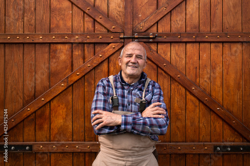 Portrait of smiling farmer with crossed arms standing by wooden barn or food granary doors at domestic animals farm.