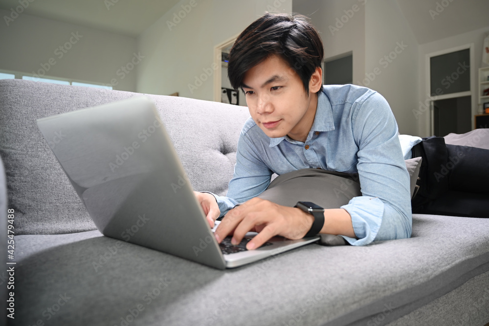 Happy young man lying on sofa in living room and surfing internet with computer laptop. Happy young man lying on sofa in living room and surfing internet with computer laptop.