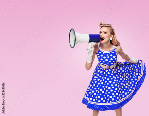 Portrait of happy excited woman holding megaphone, dressed in pin up style blue dress in polka dot white gloves, on pink background. Caucasian blond girl posing in retro fashion vintage studio image.