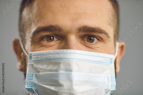 Closeup studio portrait of caucasian man in face medical mask with wide-open surprised eyes. Male face with shock emotion from coronovirus covid 19 pandemic consequences lockdown prolongation