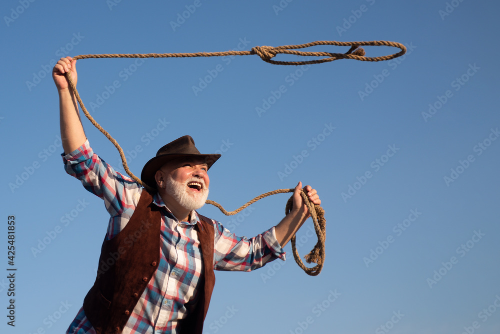 Old cowboy with lasso rope at ranch or rodeo. Bearded western man with  brown jacket and hat catching horse or cow. Stock Photo
