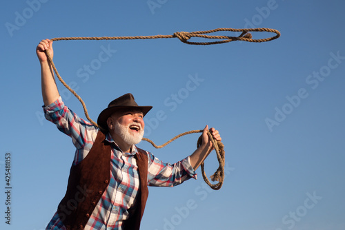 Old cowboy with lasso rope at ranch or rodeo. Bearded western man with brown jacket and hat catching horse or cow. photo