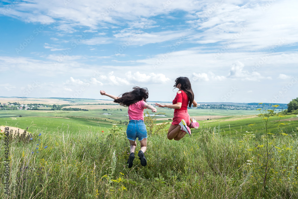 two happy slim smiling young women jumping together on rural field on the blue sky, summer time