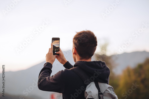 Young guy in a hoodie makes a horizontal photo of sunset on his phone in mountains