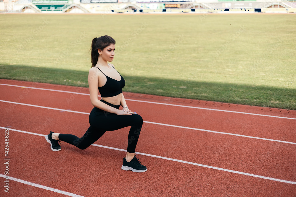 Young athletic girl stretches, prepares her body and muscles for a productive fitness workout treadmill race running track. Flexible female sporty model on the city stadium. Image with copy space.