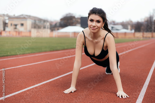 Young athletic girl doing push ups plank, prepares her body and muscles for a productive fitness workout treadmill race running track. Flexible female sporty model on the city stadium.