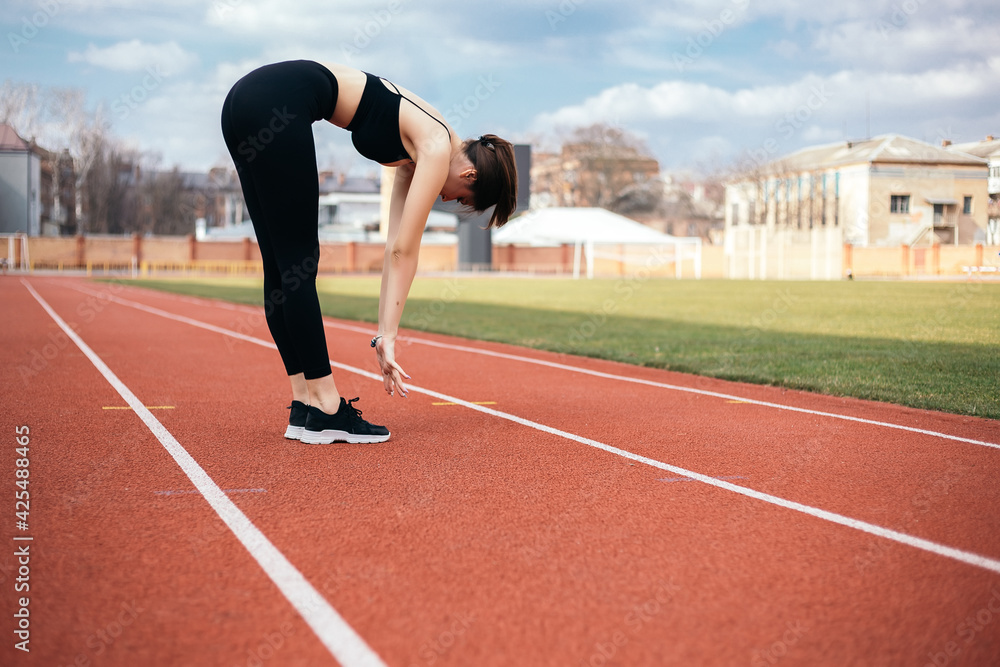 Obraz premium Young athletic girl stretches, prepares her body and muscles for a productive fitness workout treadmill race running track. Flexible female sporty model on the city stadium. Image with copy space.