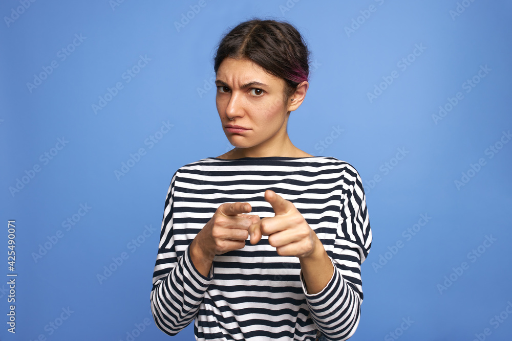 Studio shot of angry annoyed young Caucasian female in striped shirt frowning, making displeased grimace, pointing both index fingers at camera, showing warning gesture. Negative feelings and reaction Studio shot of angry annoyed young Caucasian female in striped shirt frowning, making displeased grimace, pointing both index fingers at camera, showing warning gesture. Negative feelings and reaction