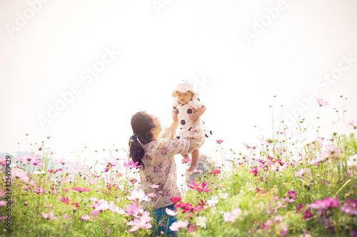 Young asian mother with her pretty curly daughter are walking in spring garden with pink blossom roses flowers, sunset time. Family holiday in summer day.