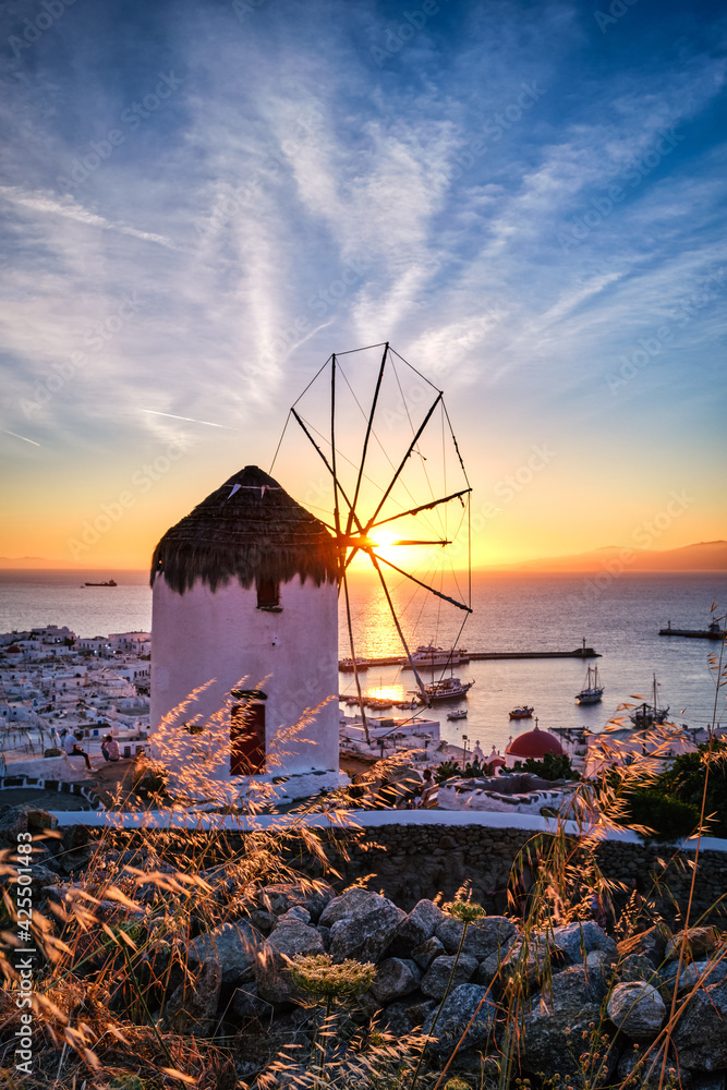 Famous traditional white windmill overlook port and harbor of Mykonos, Cyclades, Greece at sunset sky. Beautiful sky, sun, sea, horizon, town, island