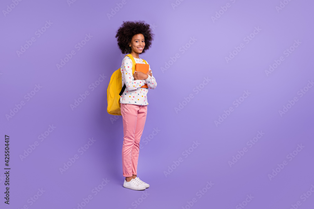 Full length profile side photo of little dark skin girl hold book wear bag pink pants school isolated on violet color background Full length profile side photo of little dark skin girl hold book wear bag pink pants school isolated on violet color background