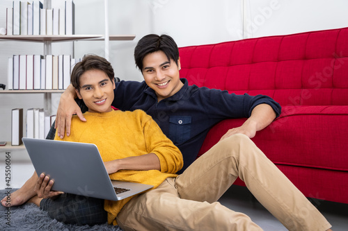 Asian gay couple sitting at sofa using laptop browsing Internet for online shopping, hand around his partner, hugging or embracing. Looking at camera.