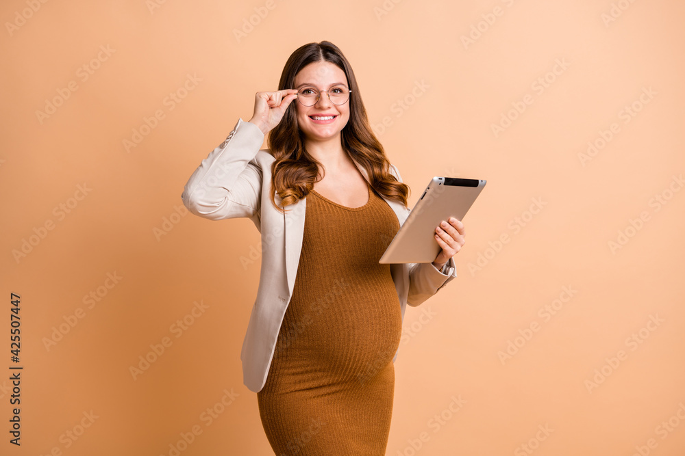 Portrait of pretty clever confident cheerful pregnant girl using ebook touching specs isolated over beige pastel color background Portrait of pretty clever confident cheerful pregnant girl using ebook touching specs isolated over beige pastel color background