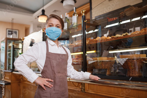 Young bakers son wearing medical mask, posing proudly at his family bakery, copy space