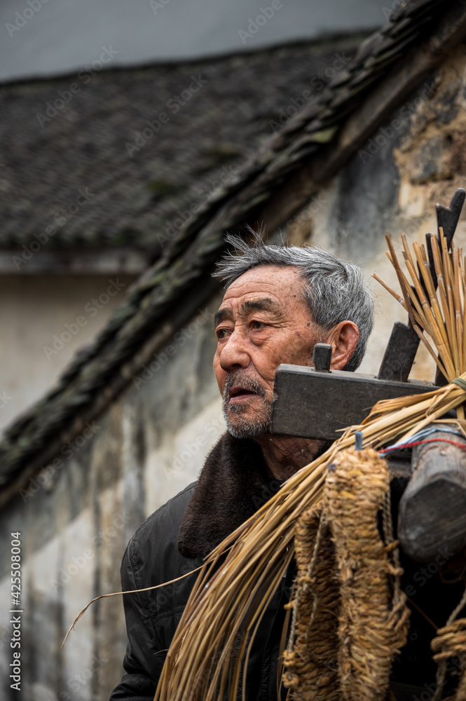 An old man stands in front of his house carrying his handcraft tools. He uses the tools to make straw sandals.