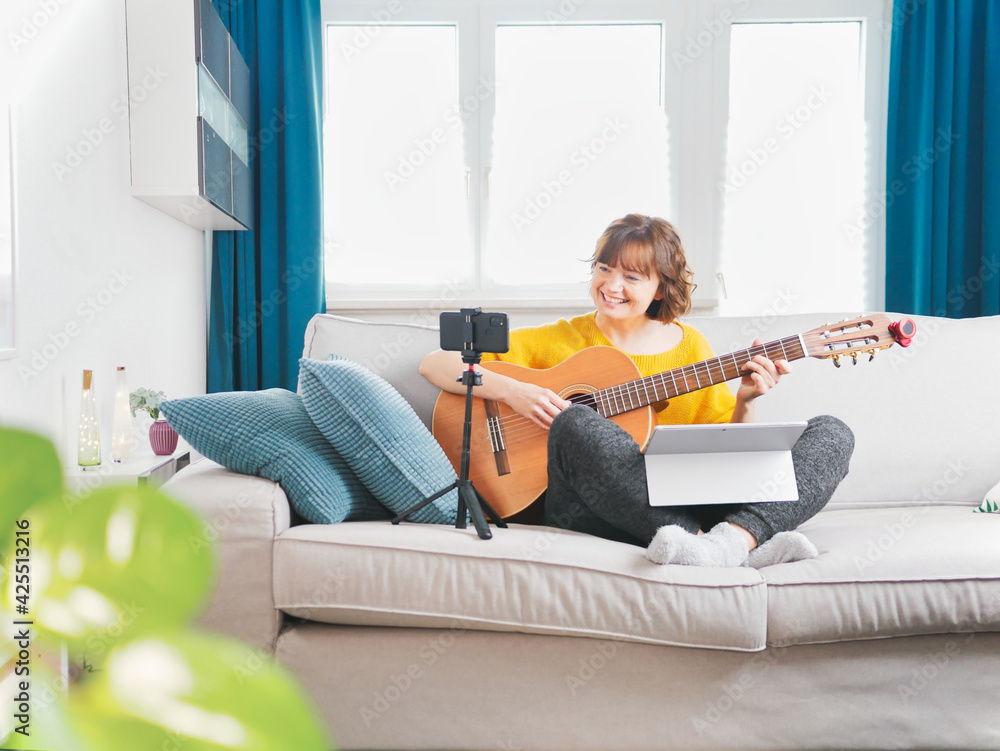 Beautiful young woman playing guitar with laptop at home. online course to learn play guitar. blogger girl sitting on sofa playing online the guitar with smartphone. coronavirus self isolation Beautiful young woman playing guitar with laptop at home. online course to learn play guitar. blogger girl sitting on sofa playing online the guitar with smartphone. coronavirus self isolation