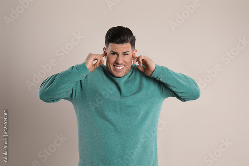 Emotional man covering ears with fingers on beige background