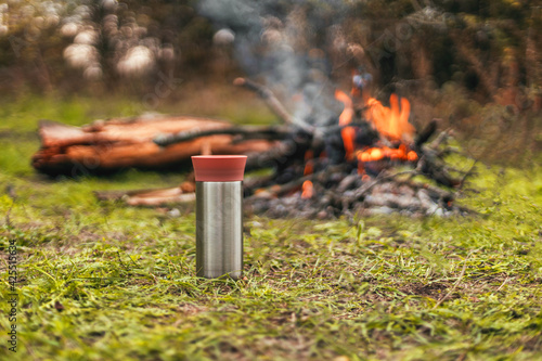 Metal thermos bottle with hot tea or coffee drink near large campfire. Trekking, adventure and seasonal vacation.