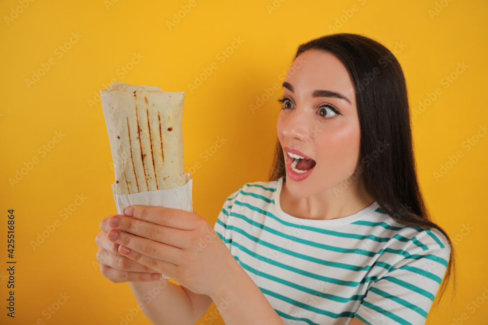 Emotional young woman holding tasty shawarma on yellow background Emotional young woman holding tasty shawarma on yellow background