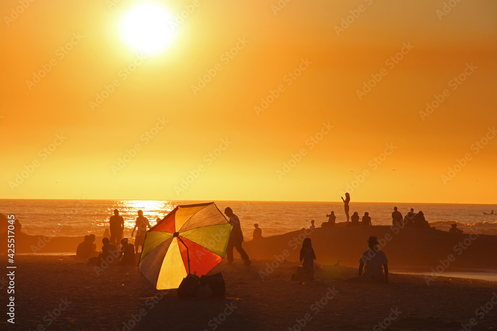 Lively summer beach scene in Cape Town, South Africa during golden hour with silhouette of people and colorful sunshade before sunset