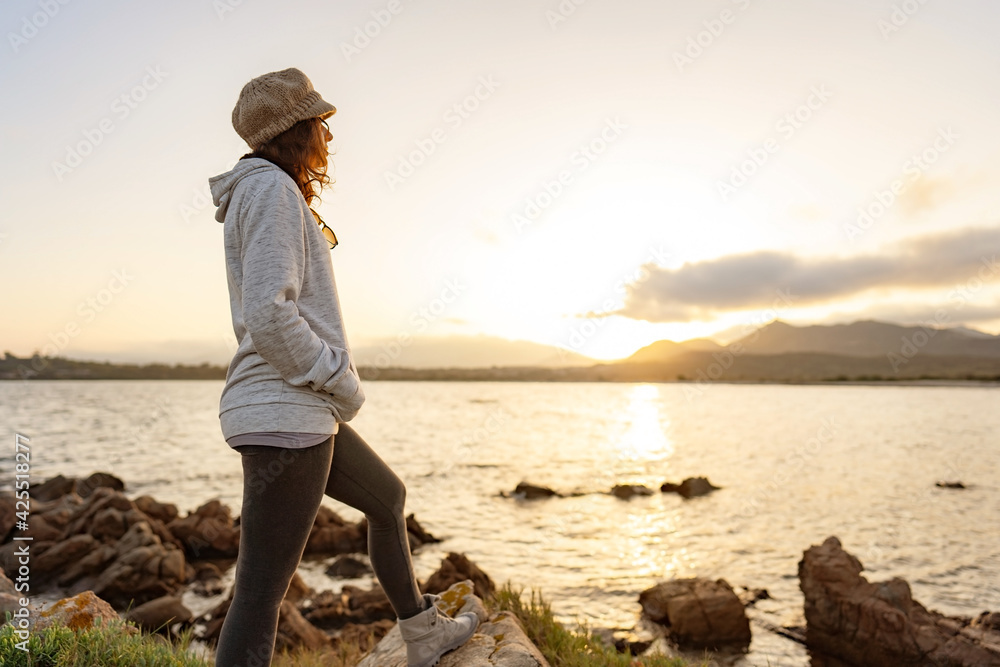 Alone pensive woman standing on sea rocks watching at sunset or sunrise between mountain on the horizon. Casual pretty person with wool hat finds her spirituality living the nature in travel vacation