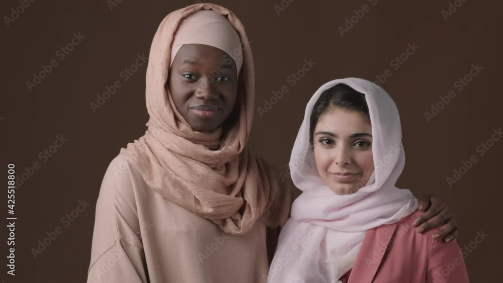 Medium shot portrait of young Muslim mixed-race and African-American women wearing hijab and abaya standing together against brown background and looking at camera Medium shot portrait of young Muslim mixed-race and African-American women wearing hijab and abaya standing together against brown background and looking at camera