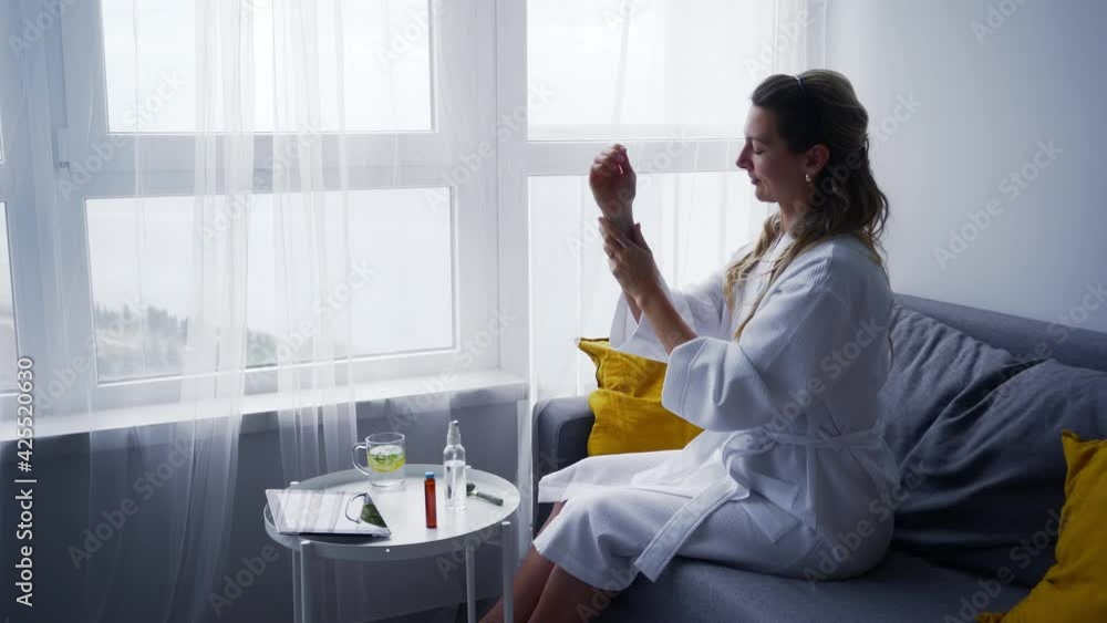 Attractive calm young woman in white tidy bathrobe applies coconut butter on hands sitting on comfortable sofa near large window in living room Attractive calm young woman in white tidy bathrobe applies coconut butter on hands sitting on comfortable sofa near large window in living room