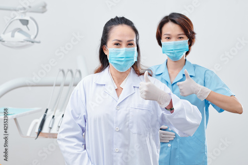 Portrait of cheerful experienced dentist and nurse in protective masks and silicone gloves showing thumbs-up and looking at camera