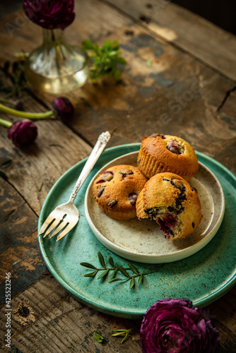 Three homemade cherry and chocolate muffins and ranunculus flowers on ceramic plates on old rustic wooden table. 