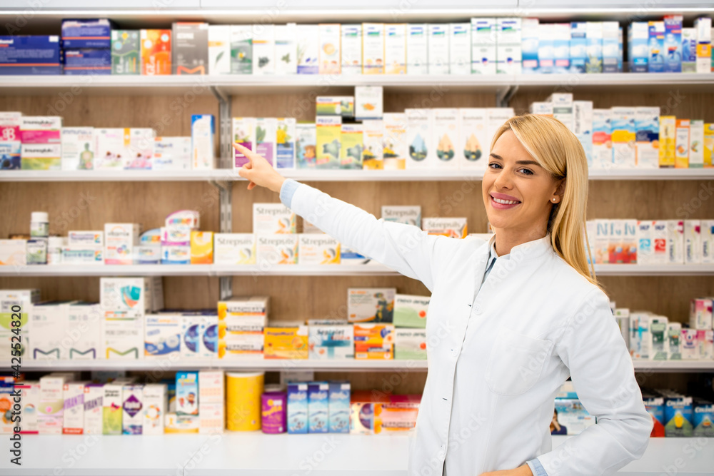 Portrait of smiling female caucasian pharmacist standing in drug store pointing at the shelf.