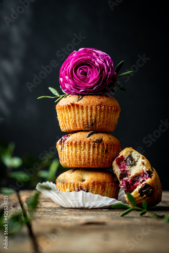 Stack of three homemade cherry and chocolate muffins with ranunculus flower on top and bitten one on black background with shadows.