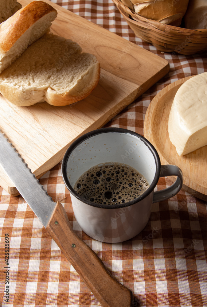 Breakfast table in Brazil with breads, cheese, cup of coffee and accessories on a brown and beige checkered tablecloth, dark background, selective focus. Breakfast table in Brazil with breads, cheese, cup of coffee and accessories on a brown and beige checkered tablecloth, dark background, selective focus.