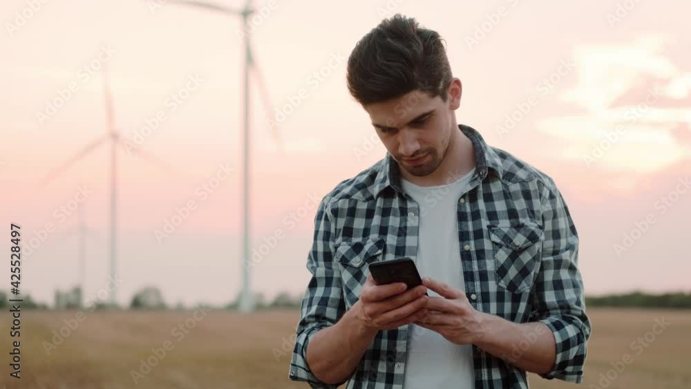 A focused man wearing shirt is using his smartphone standing at the electric windmill farm A focused man wearing shirt is using his smartphone standing at the electric windmill farm