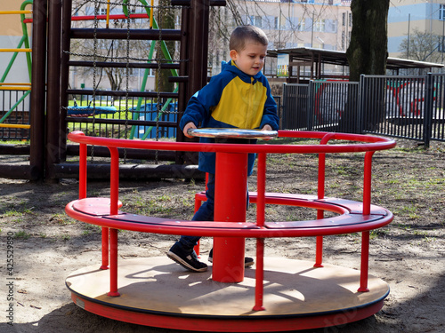 a bright boy in a blue and yellow jacket rides on a red round swing on a sunny spring day at the playground . outdoor activities